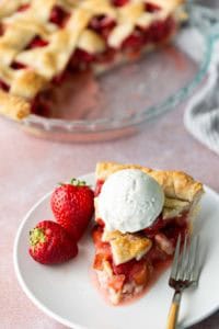 A slice of strawberry pie topped with a scoop of vanilla ice cream, served on a plate with two fresh strawberries and a fork. The rest of the pie is in the background.