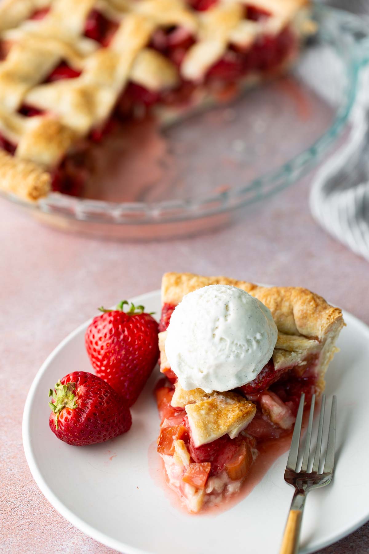 A slice of strawberry pie topped with a scoop of vanilla ice cream, served on a plate with two fresh strawberries and a fork. The rest of the pie is in the background.