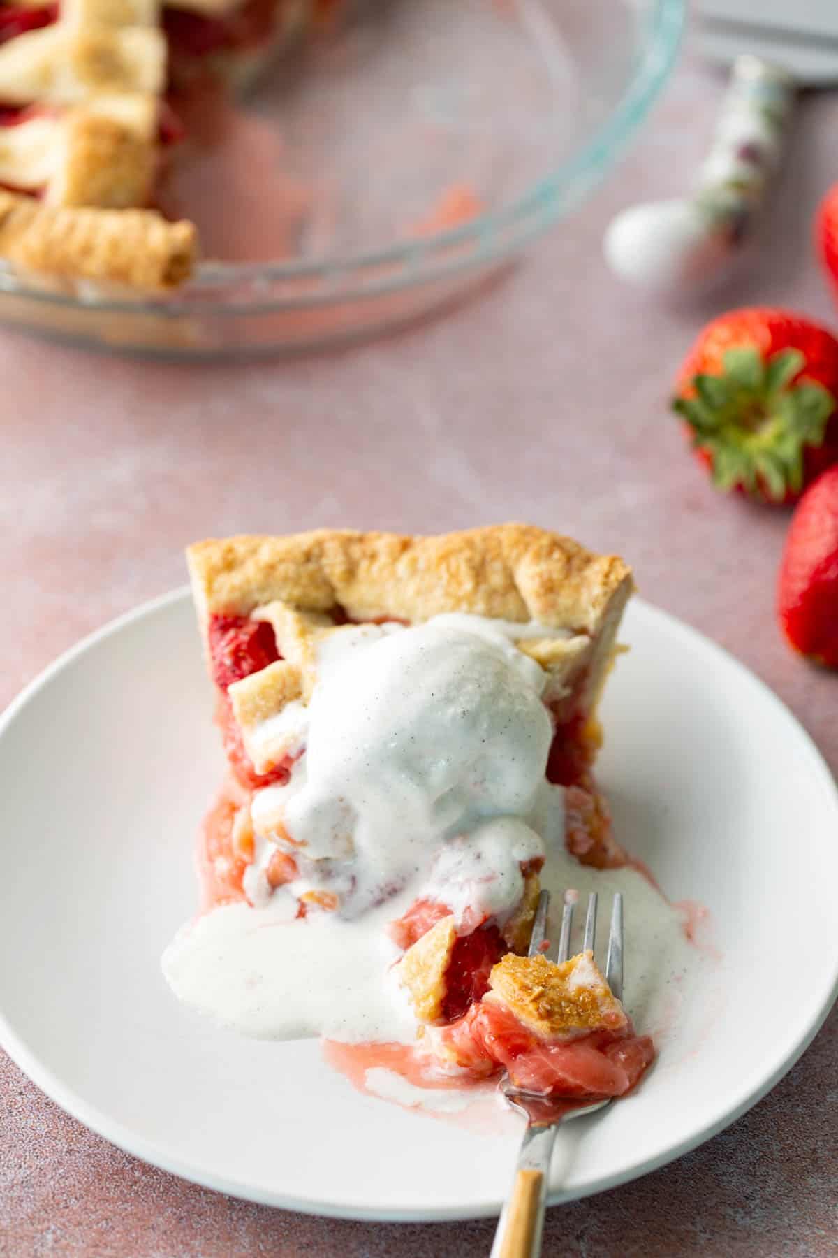 A slice of strawberry pie topped with melting vanilla ice cream sits on a white plate with a fork; whole strawberries and the pie dish are in the background.