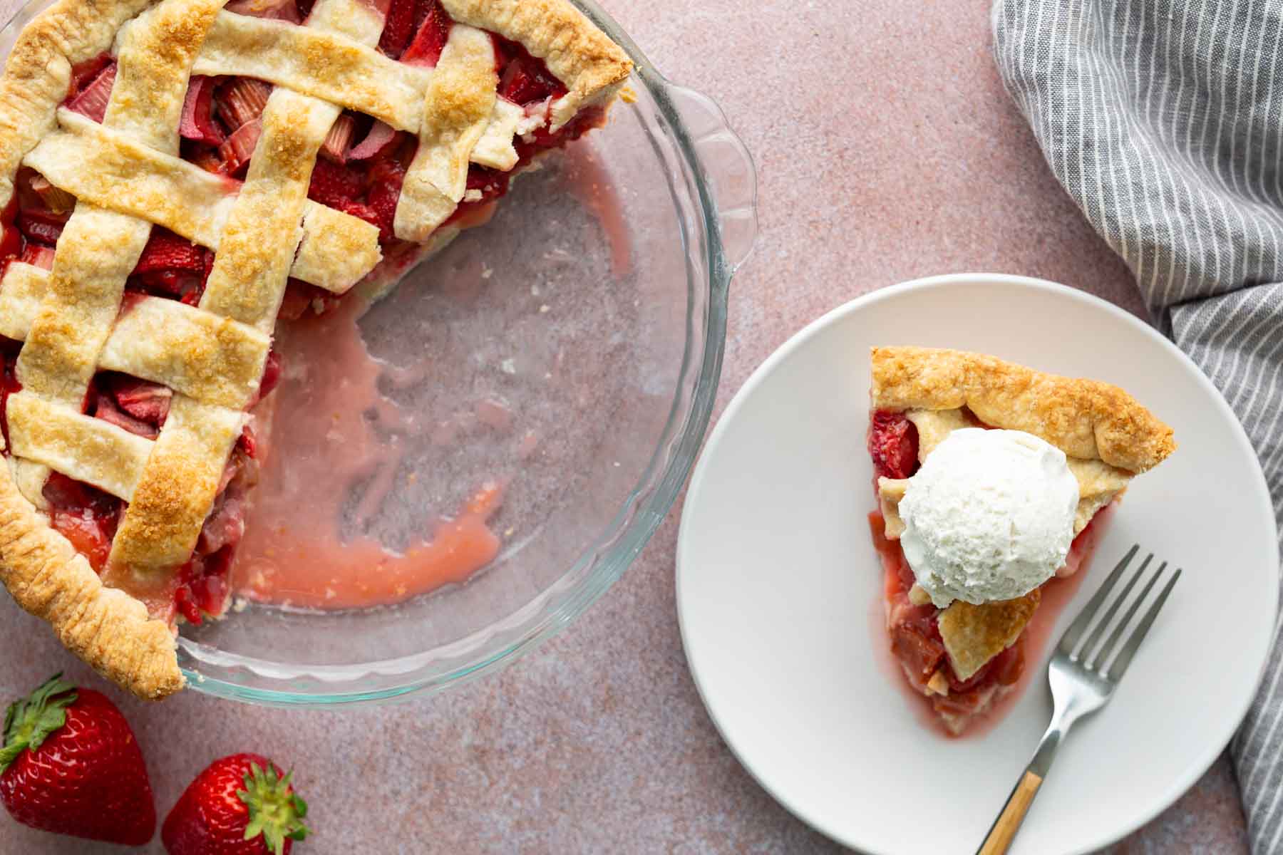 A partially eaten strawberry pie in a glass dish next to a plate with a slice of pie topped with a scoop of vanilla ice cream and a fork. Two strawberries and a striped napkin are nearby.
