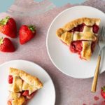 Two slices of strawberry rhubarb pie on white plates with a fork, next to fresh strawberries on a pink surface.