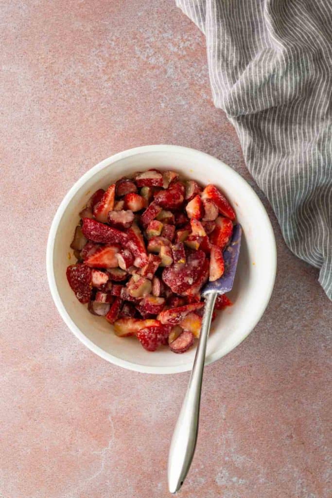A white bowl filled with chopped strawberries and rhubarb, mixed with a spatula, sits on a pink surface next to a gray striped cloth.