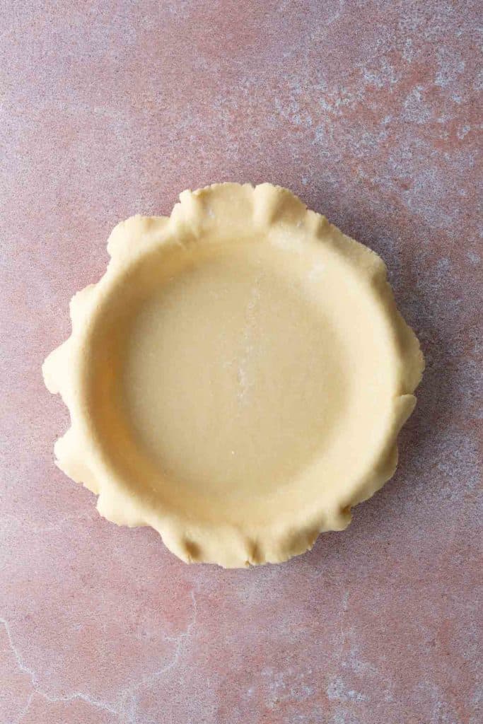 Unbaked pie crust in a round pan with crimped edges sits on a light pink textured surface.