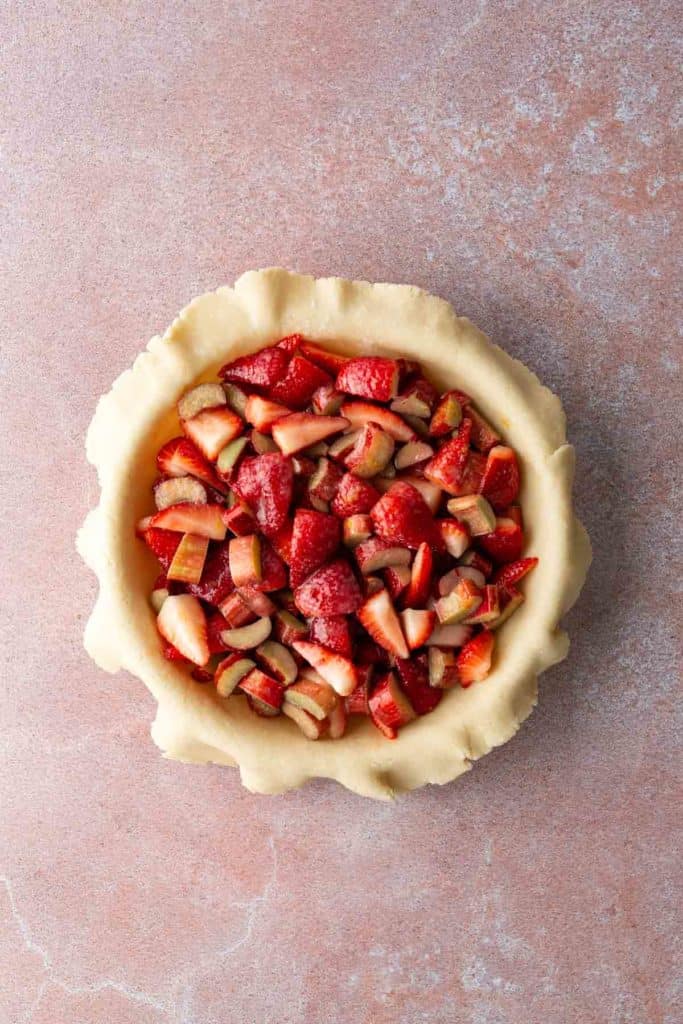 Overhead view of an unbaked pie crust filled with chopped strawberries and rhubarb on a light pink surface.
