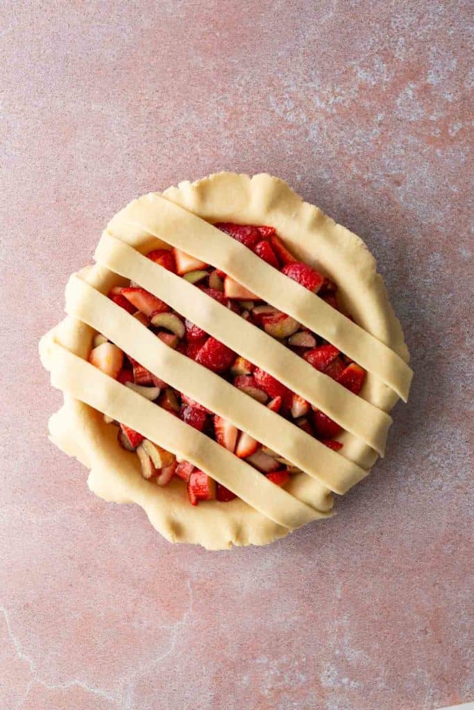 Unbaked pie with a fruit filling and lattice pastry strips on top, set on a light pink textured surface.