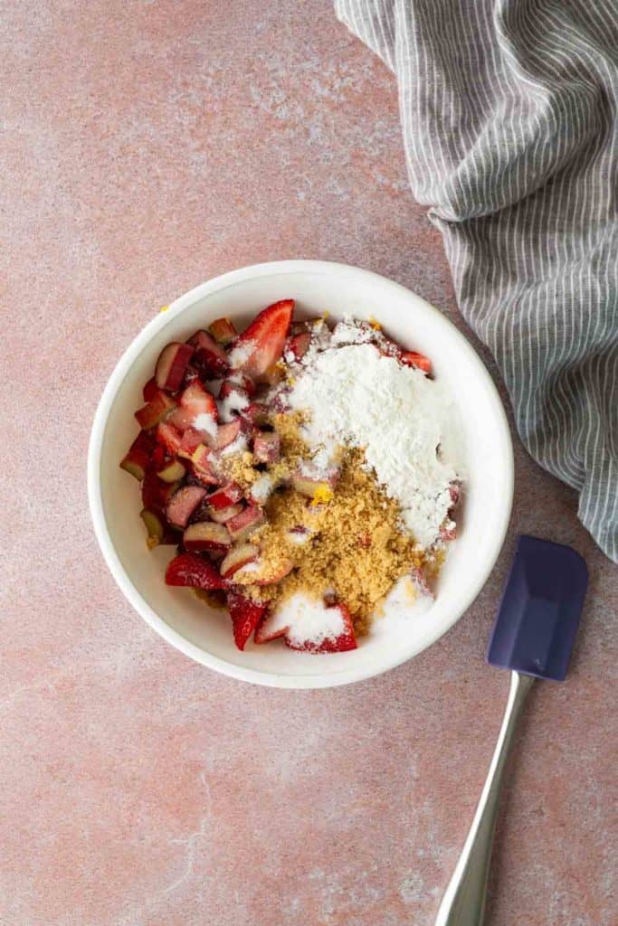 A white bowl containing sliced strawberries, brown sugar, white sugar, and flour sits on a pink surface next to a blue spatula and a gray striped cloth.