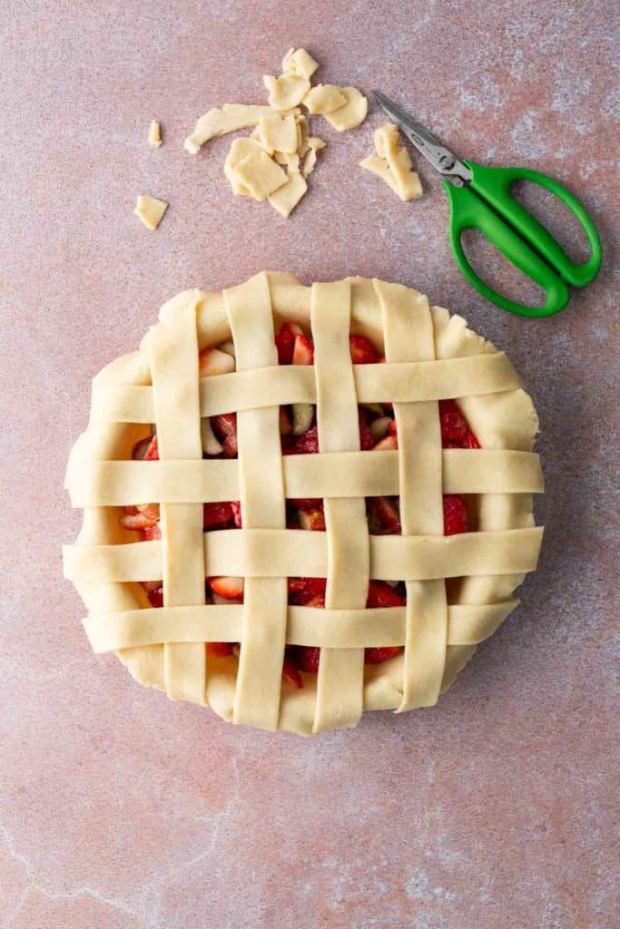 Unbaked lattice-top pie with raw dough strips on top, some trimmed pastry scraps, and a green-handled pair of scissors on a pink surface.