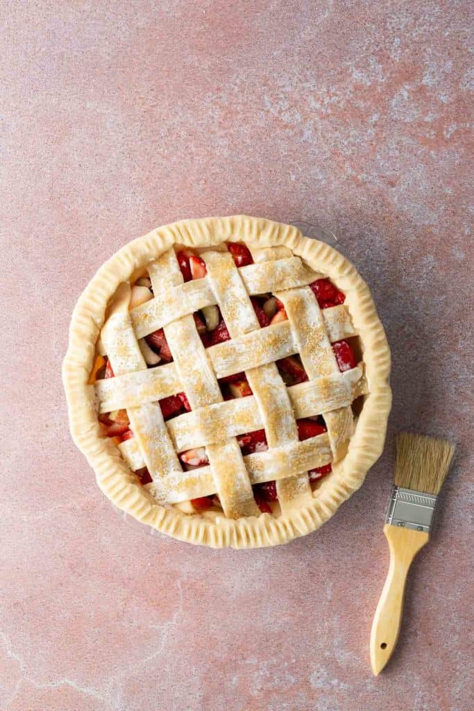 A lattice-topped, unbaked fruit pie sits on a pink surface next to a pastry brush.