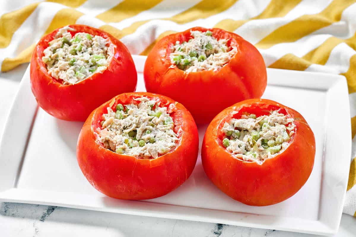Four hollowed-out tomatoes filled with a mixture of tuna salad and chopped green vegetables, arranged on a rectangular white plate.