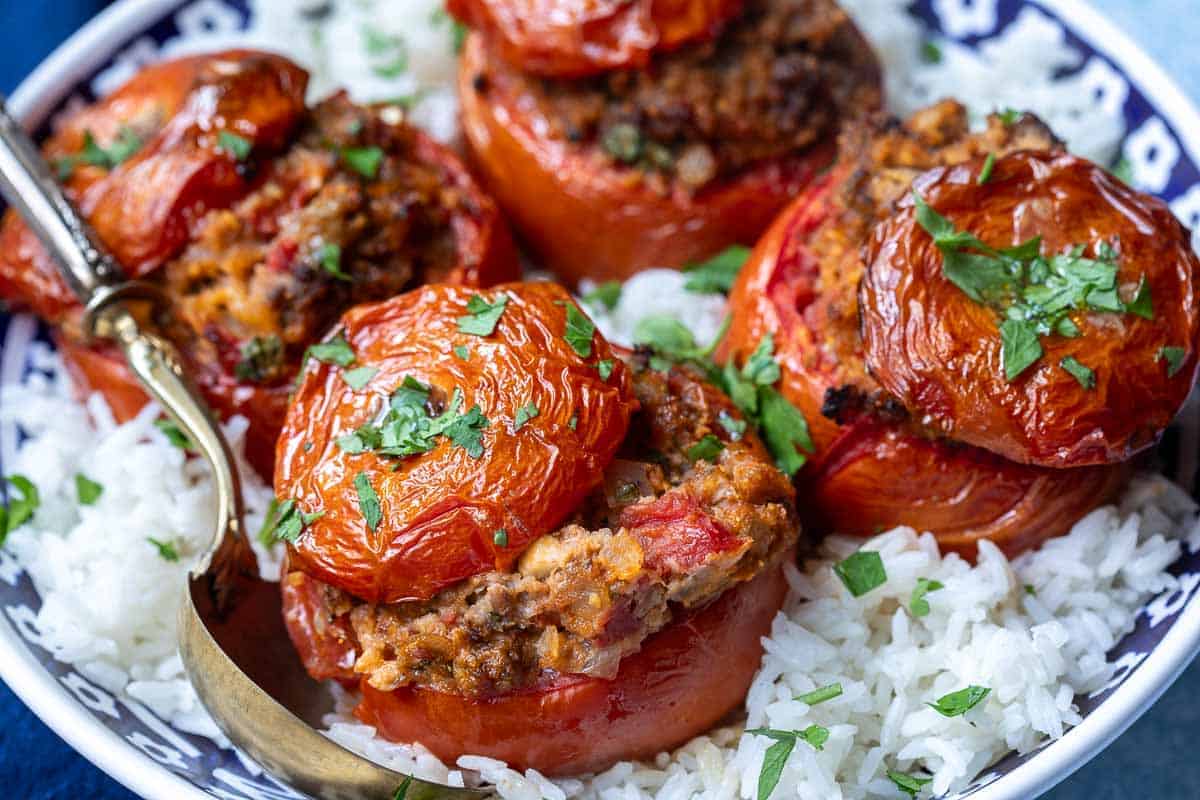 Four baked stuffed tomatoes filled with a meat and herb mixture, garnished with chopped parsley, served over a bed of white rice on a patterned plate.