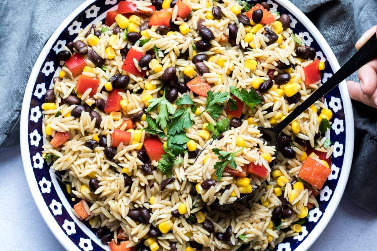 A bowl of orzo salad with black beans, corn, red bell peppers, and fresh cilantro, with a spoon scooping some from the bowl.