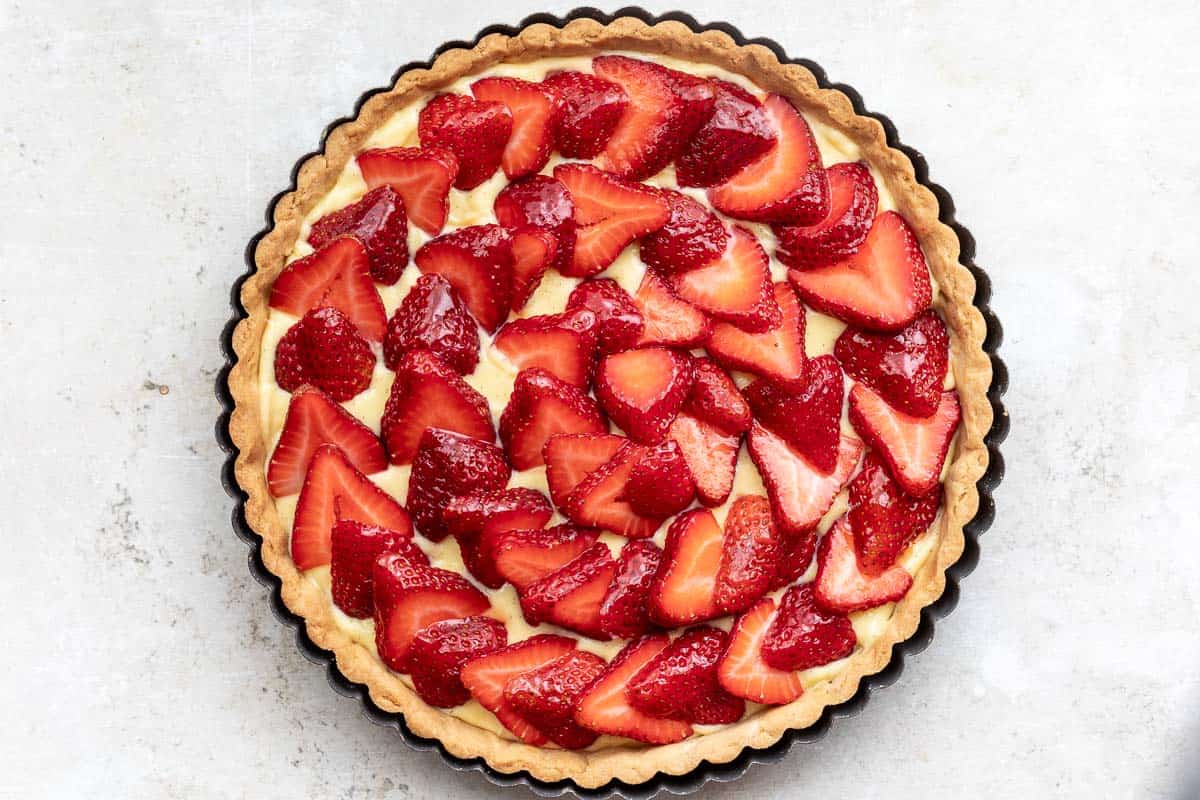 A round tart with a golden crust, filled with cream and topped with sliced fresh strawberries, arranged in a circular pattern.