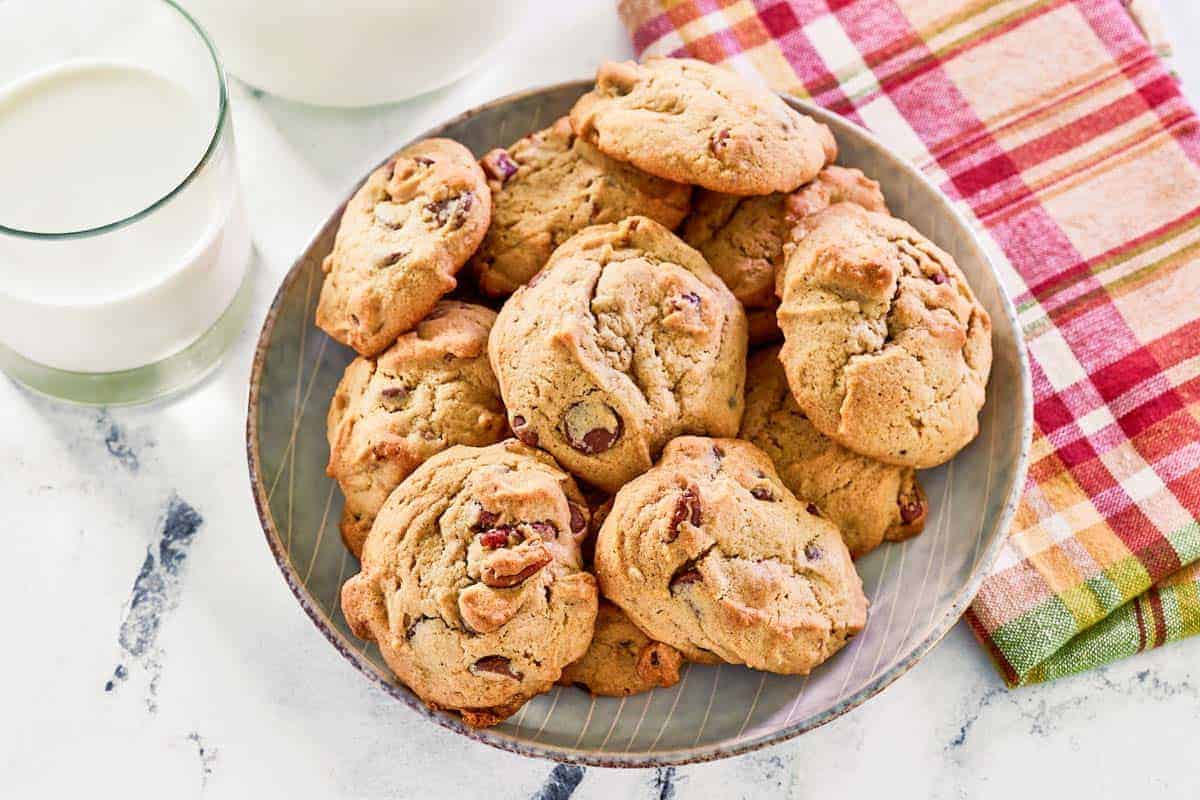 A plate of chocolate chip cookies next to a glass of milk and a colorful plaid napkin on a white marble surface.