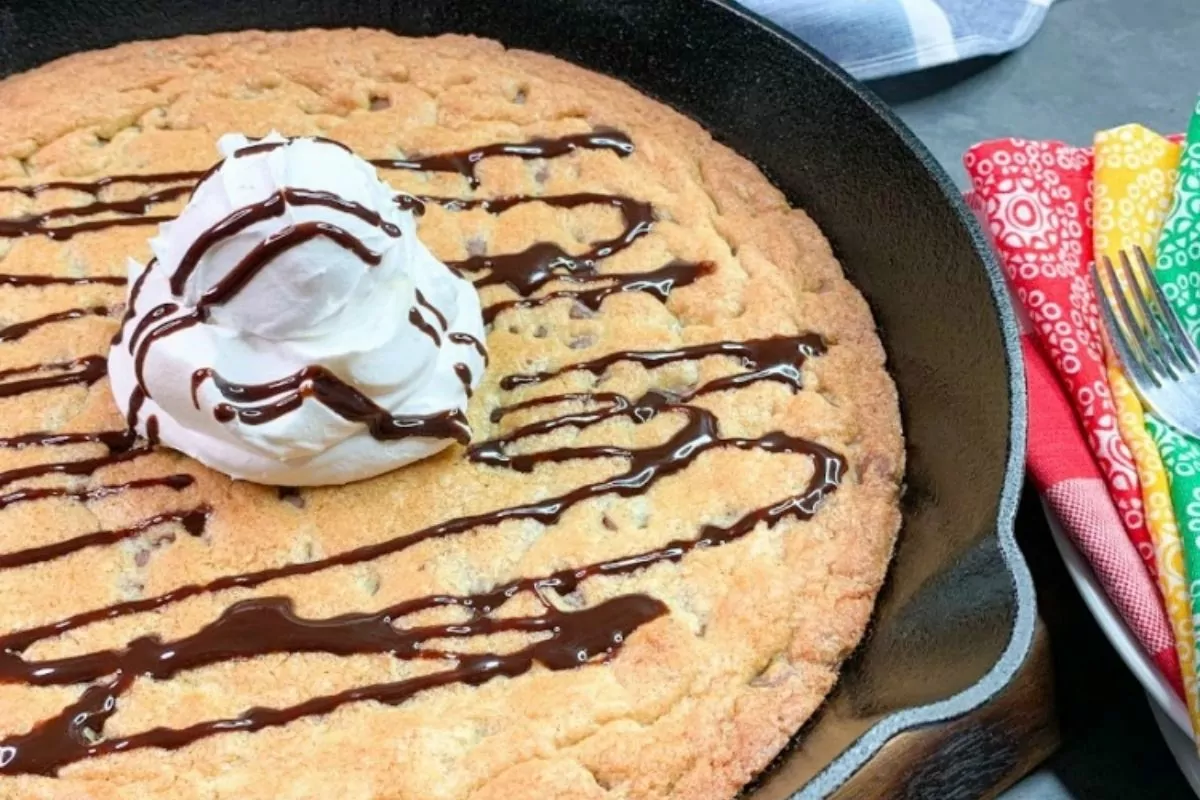 A large chocolate chip cookie in a cast iron skillet, topped with a swirl of whipped cream and drizzled with chocolate sauce. Forks and colorful napkins are beside the skillet.