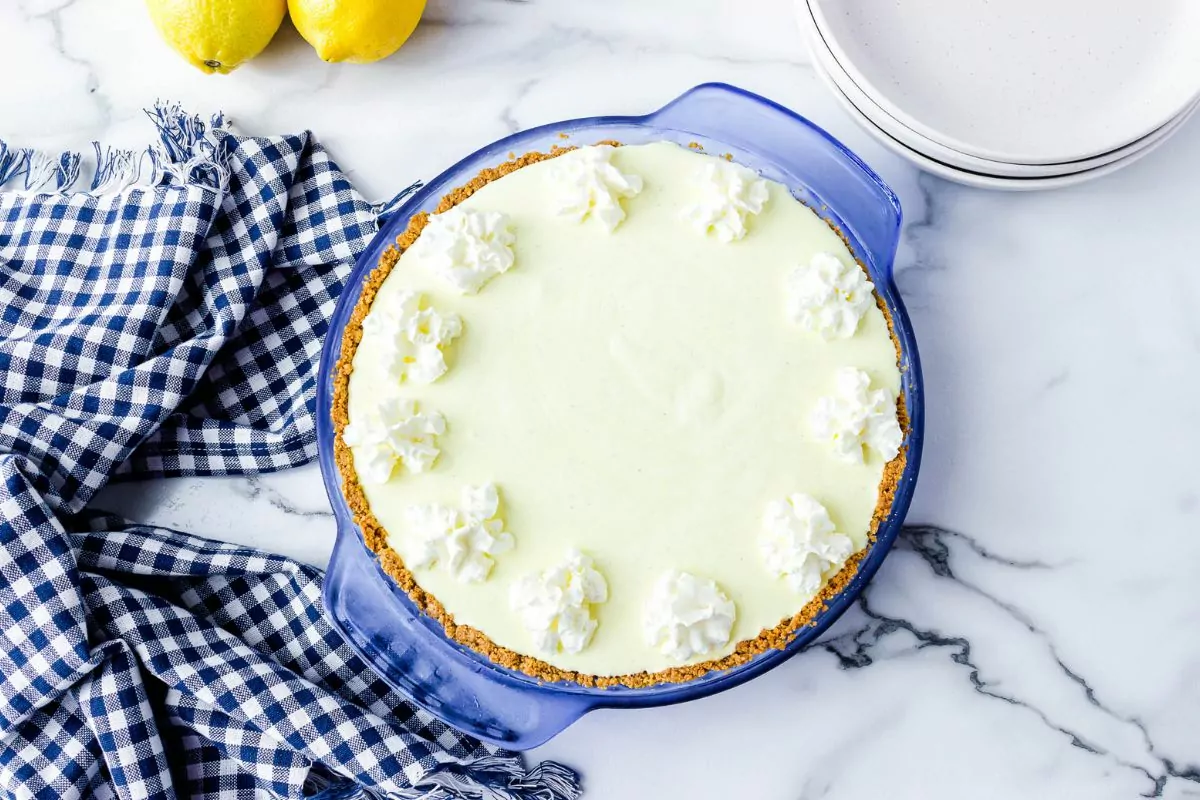 A lemon pie with a graham cracker crust and whipped cream dollops sits in a blue glass pie dish on a marble surface with lemons, plates, and a blue checked cloth nearby.