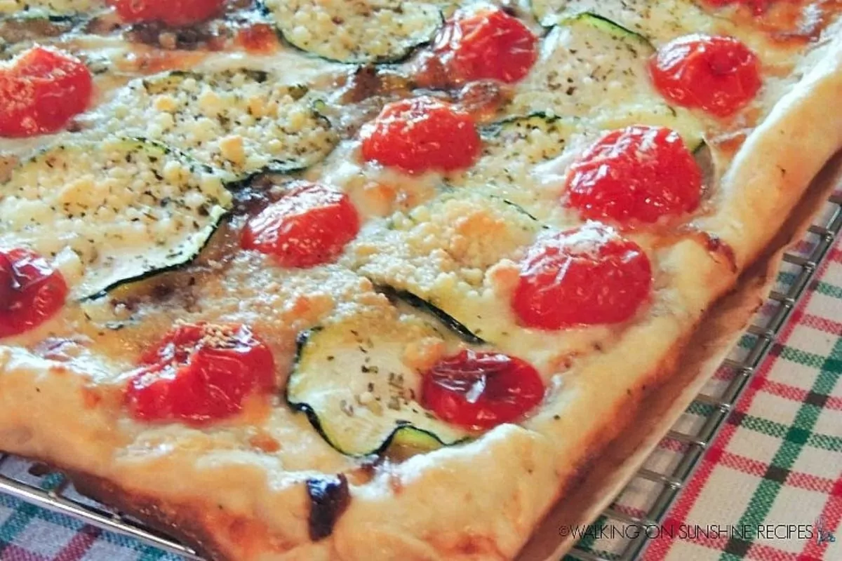 Rectangular pizza topped with sliced zucchini, cherry tomatoes, grated cheese, and herbs on a wire cooling rack over a checkered tablecloth.