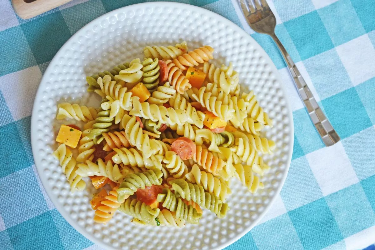 A white plate with tri-color rotini pasta salad containing cheese cubes and sliced vegetables on a blue and white checkered tablecloth.