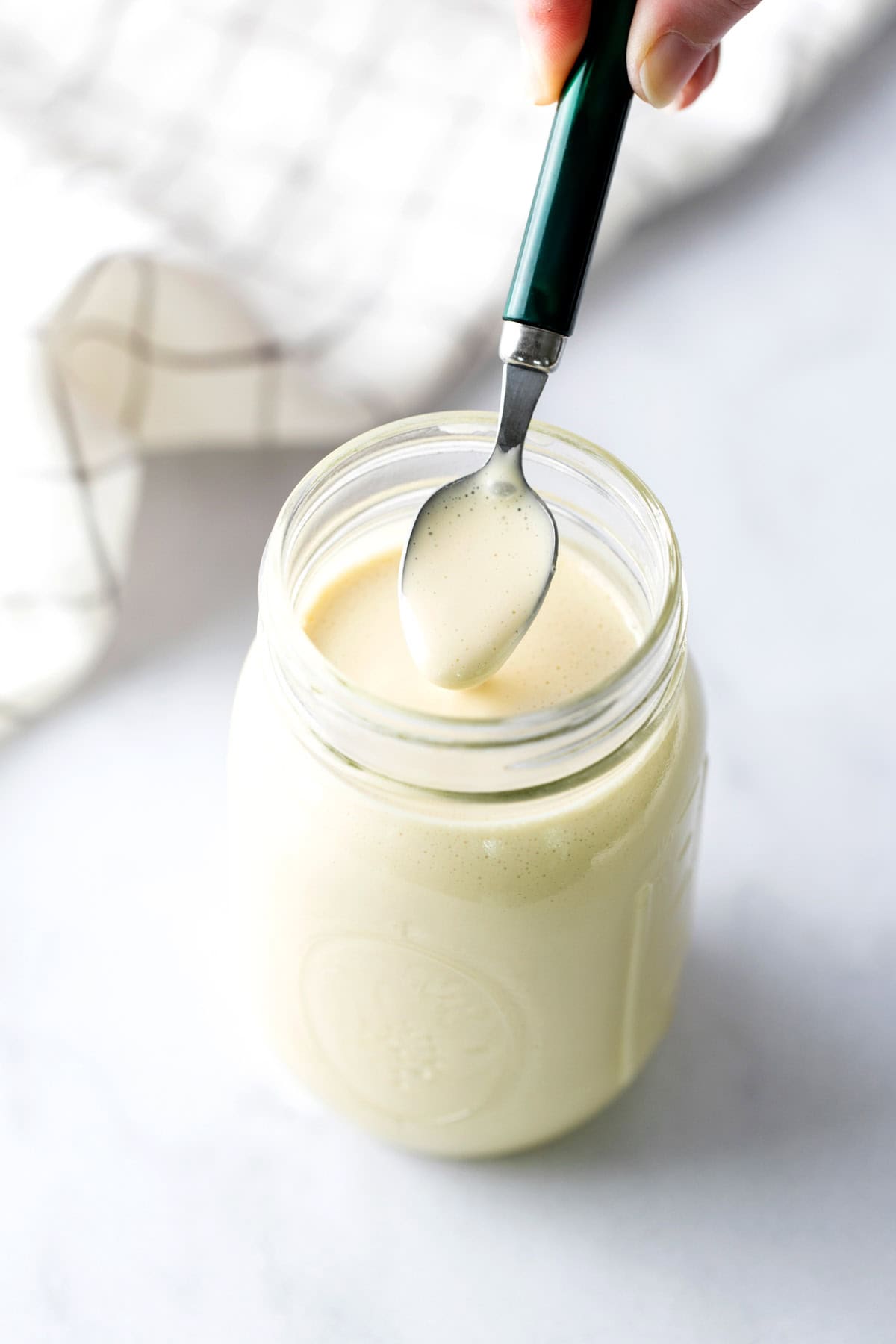 A hand holds a spoon above a jar filled with creamy white sauce or dressing on a light surface, with a checkered cloth in the background.