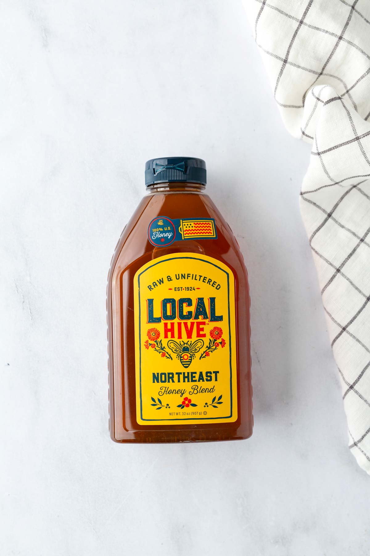 A bottle of Local Hive Northeast Honey Blend with a yellow label sits on a white surface next to a folded checkered cloth.