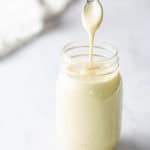 A spoon is held above a glass jar filled with a thick, creamy liquid, with some of the liquid dripping from the spoon.