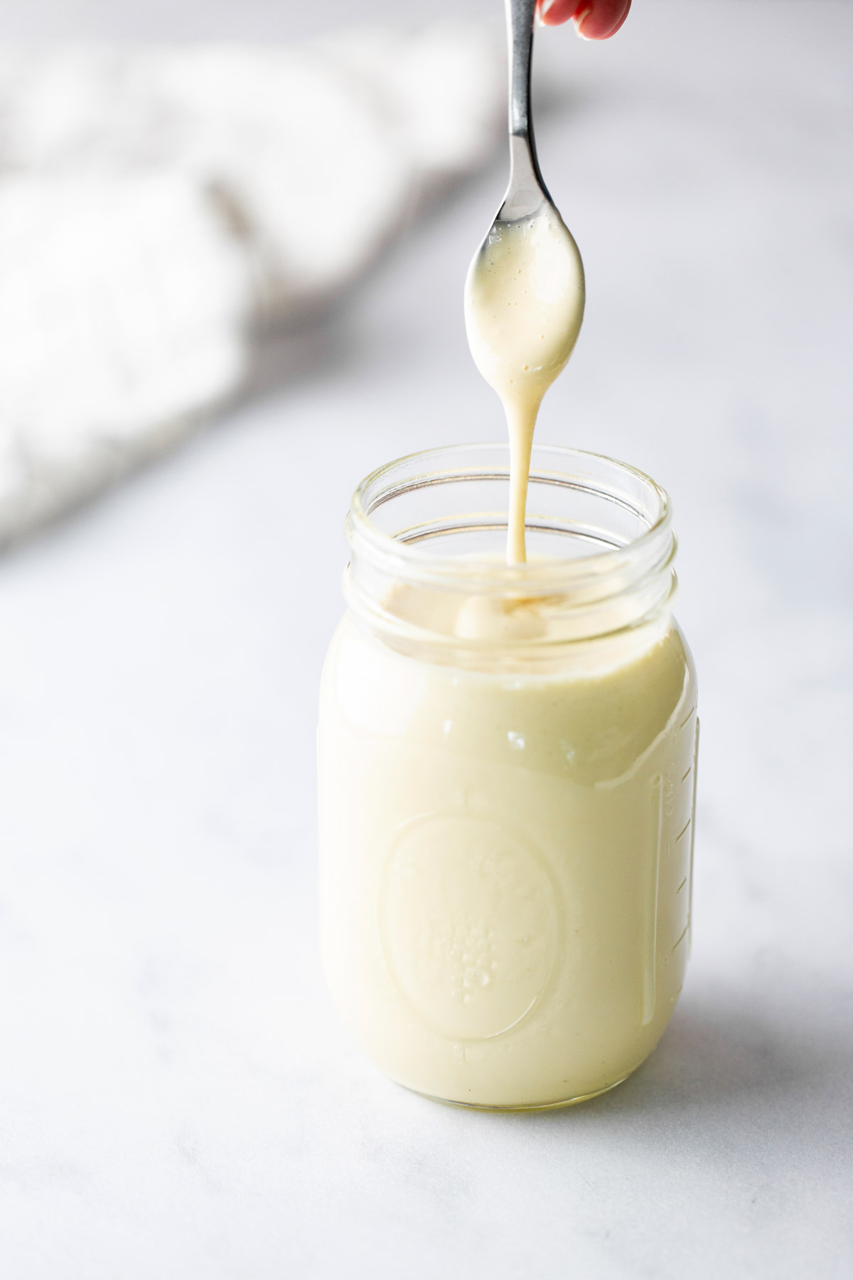 A spoon is held above a glass jar filled with a thick, creamy liquid, with some of the liquid dripping from the spoon.