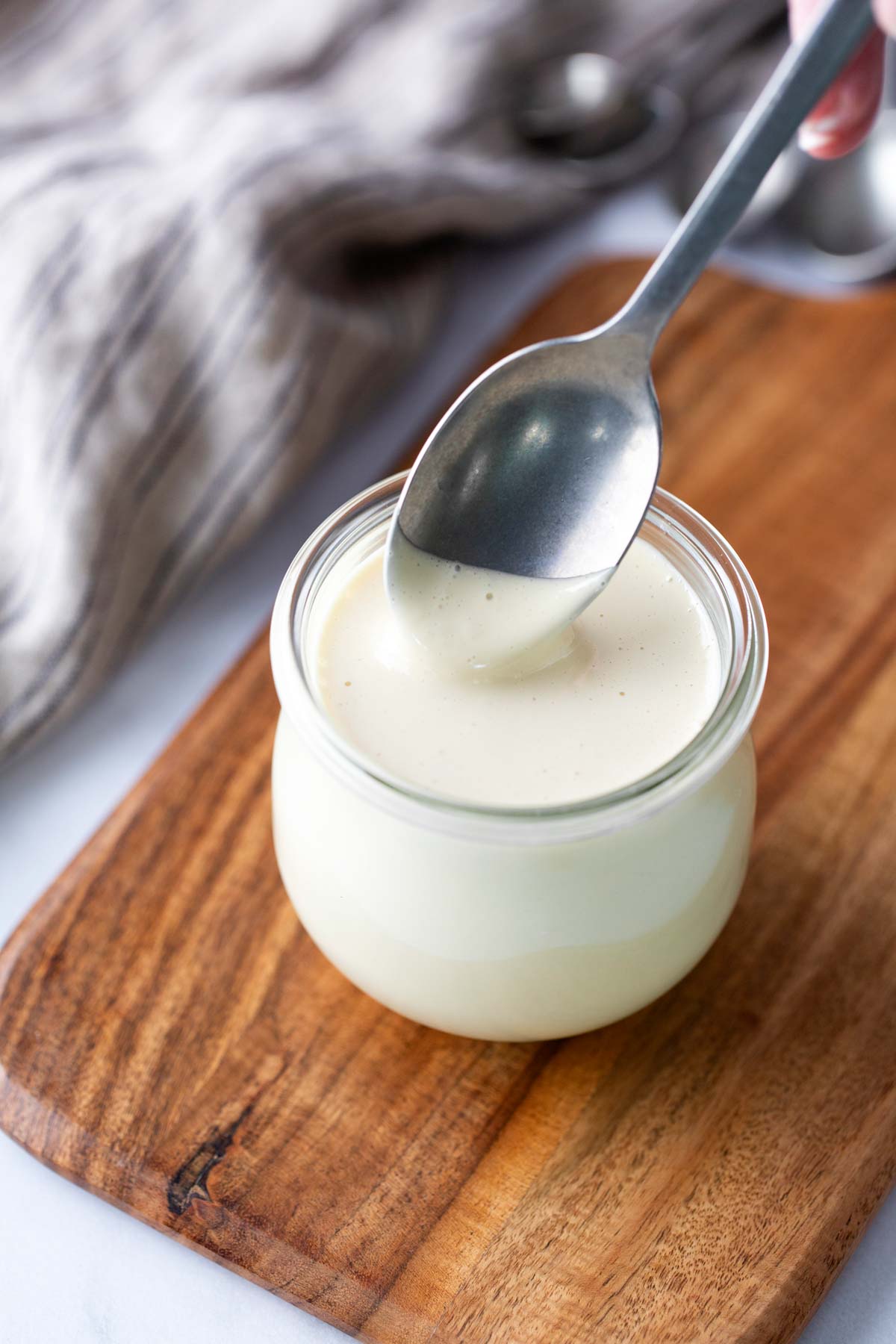 A spoon is being dipped into a small glass jar filled with a creamy white sauce, placed on a wooden board.
