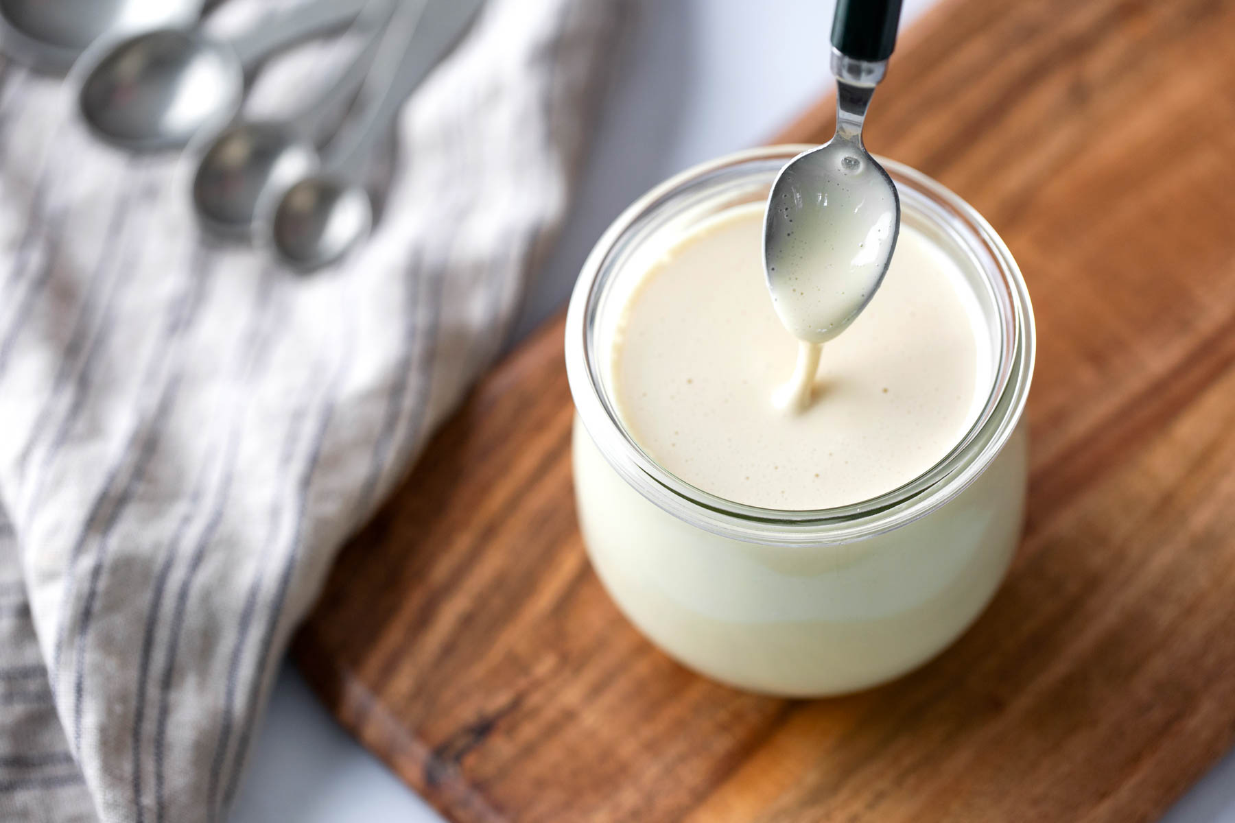A spoon is dipped into a small glass jar filled with creamy white sauce, placed on a wooden board next to a striped cloth and metal measuring spoons.