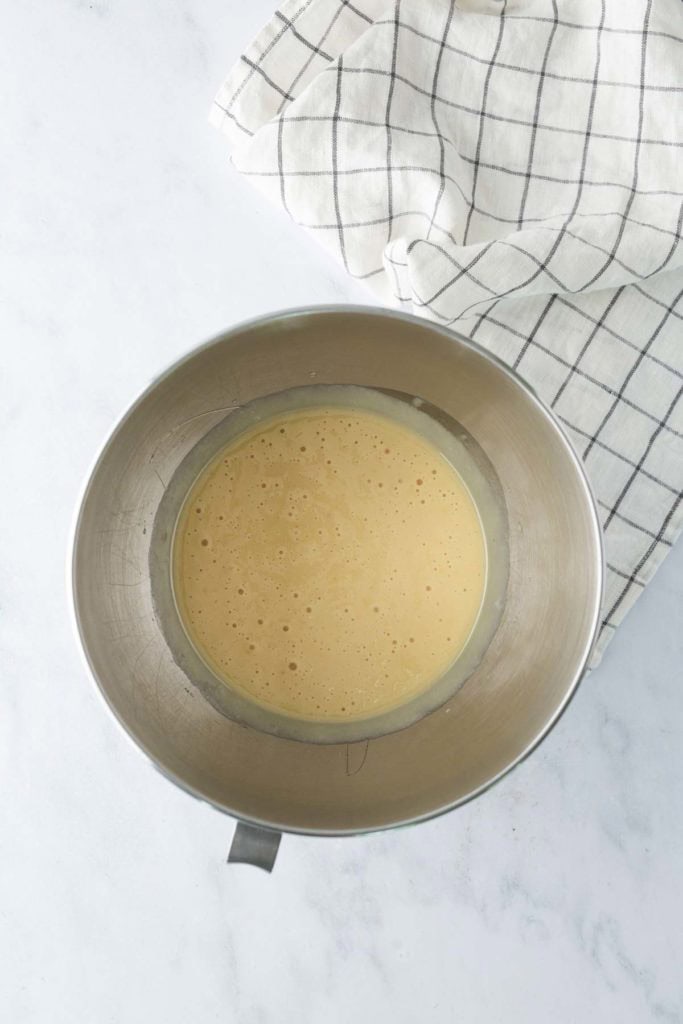 A metal mixing bowl with light yellow batter sits on a white surface next to a white kitchen towel with a black grid pattern.
