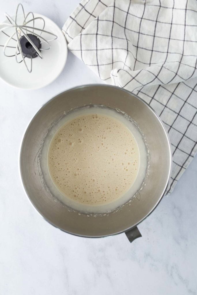 A metal mixing bowl filled with a light, frothy batter sits on a white surface next to a whisk and a folded checkered kitchen towel.