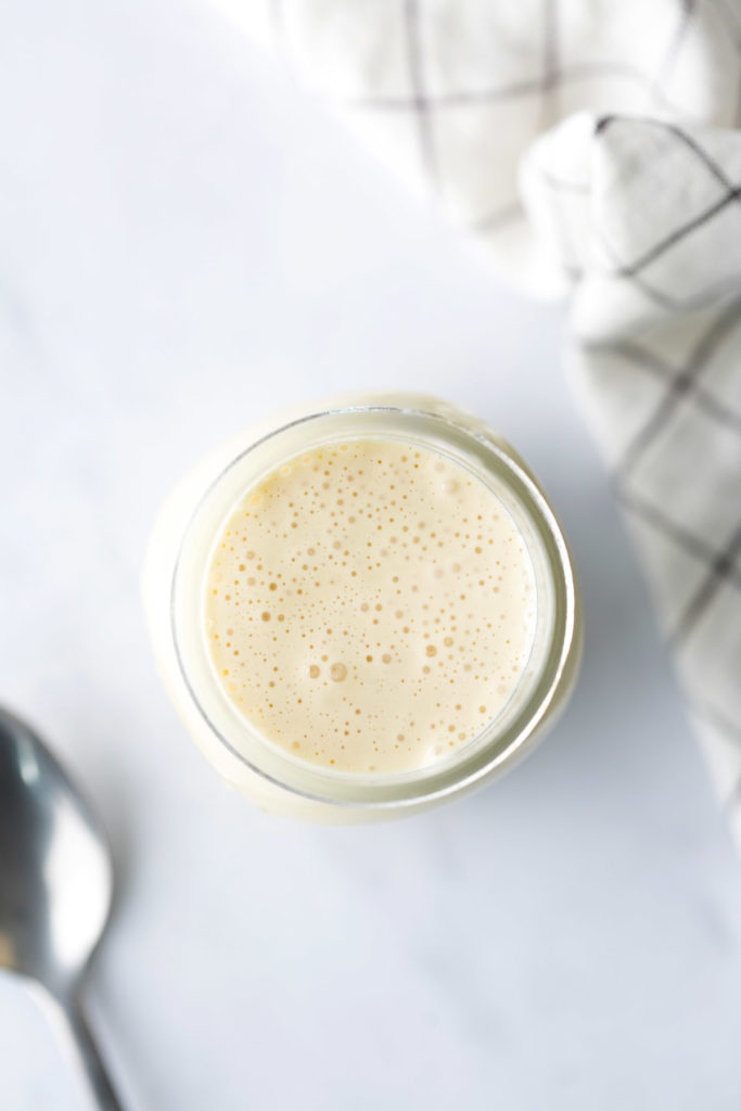 A glass jar filled with a creamy, light-colored liquid sits on a white surface next to a metal spoon and a white cloth with a black grid pattern.