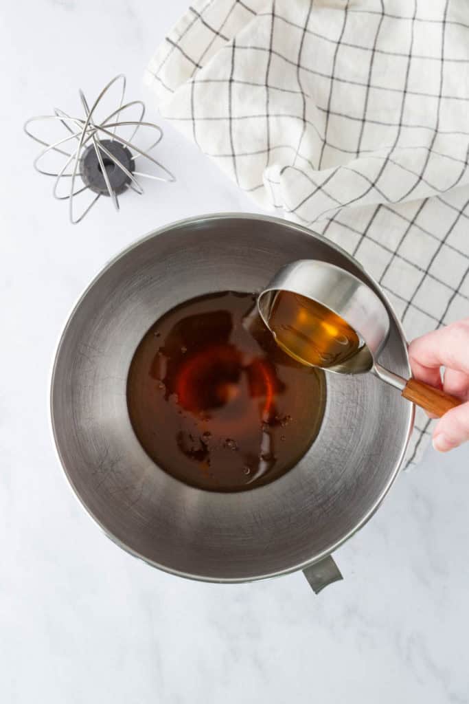 A hand pours brown liquid from a measuring cup into a metal mixing bowl on a countertop, with a whisk and checkered cloth nearby.