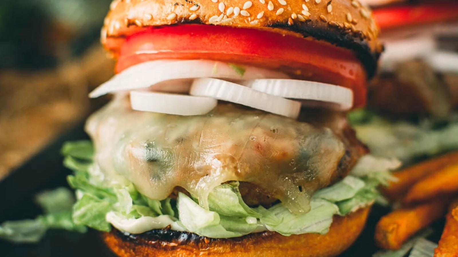 Close-up of a cheeseburger with lettuce, sliced onions, tomato, melted cheese, and a sesame seed bun, next to French fries.