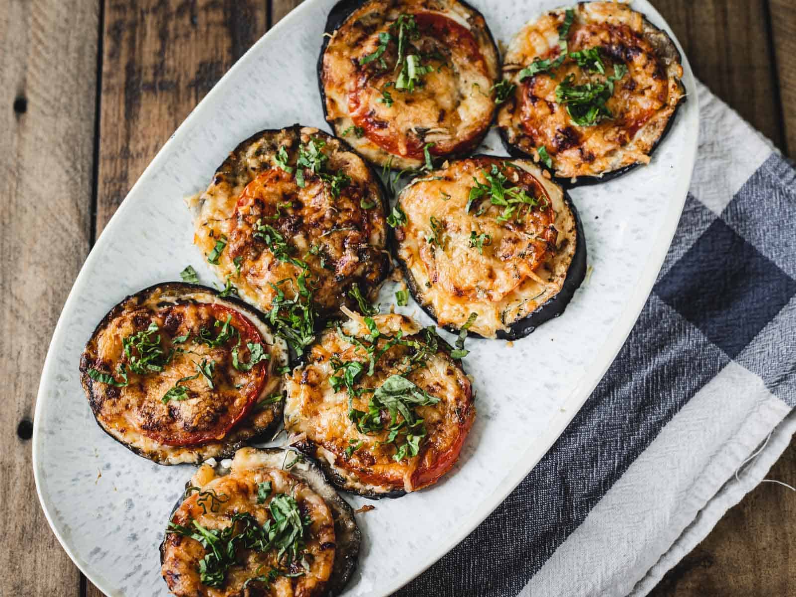 Grilled eggplant slices topped with melted cheese, tomato, and fresh herbs arranged on a white oval plate.