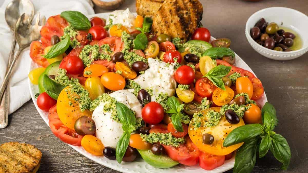 A platter of colorful sliced tomatoes, mozzarella, olives, fresh basil, pesto, and toasted bread on a table.