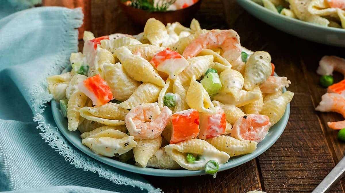 A plate of creamy pasta shells mixed with shrimp, imitation crab, green peas, and celery, served on a wooden table next to a light blue napkin.