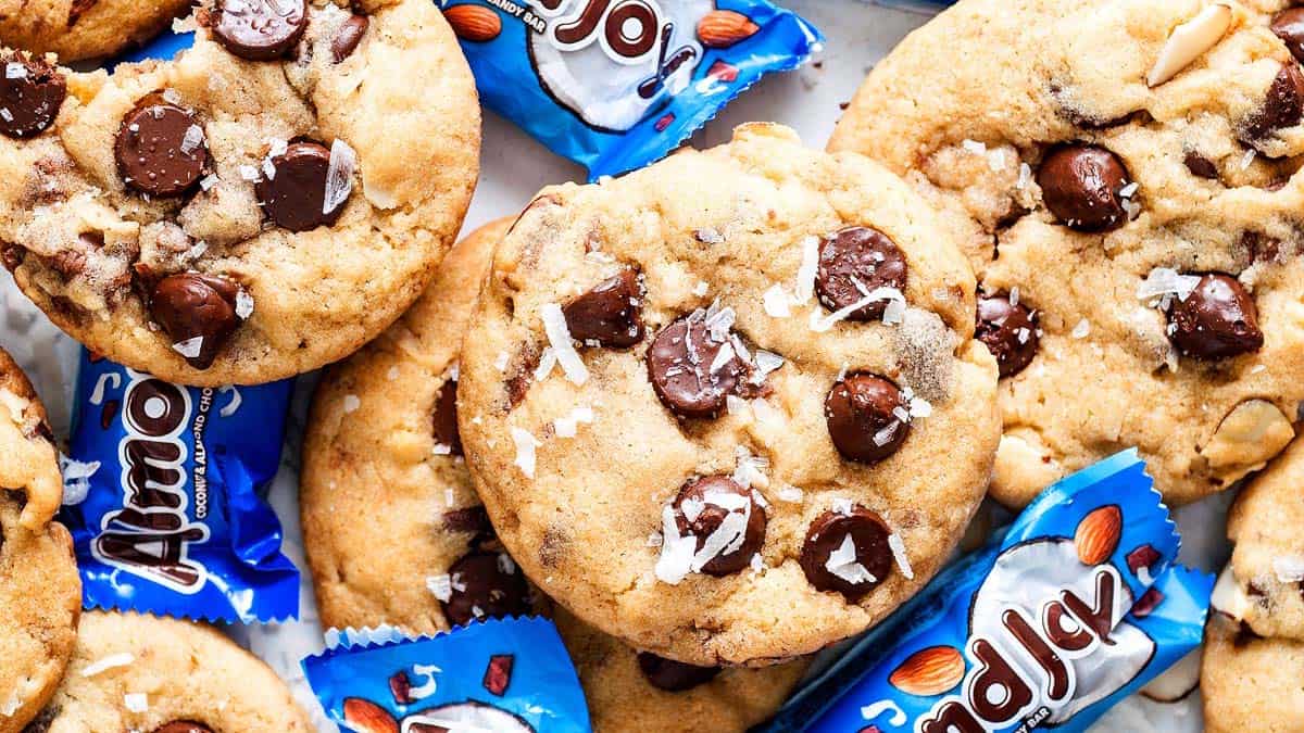 Chocolate chip cookies with sea salt are arranged on a surface alongside several unwrapped Almond Joy candy bars in blue packaging.