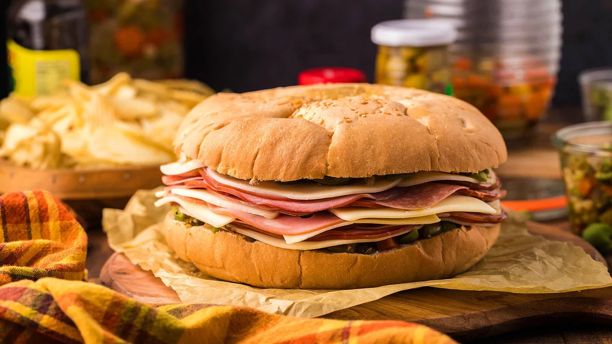 A large sandwich with layers of deli meats and cheese on a round sesame bun, placed on parchment paper with chips and jars of pickled vegetables in the background.