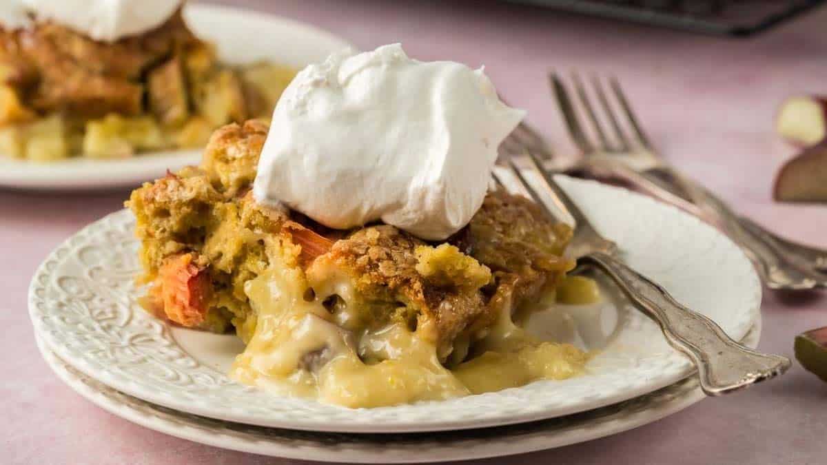 A slice of fruit cobbler topped with whipped cream on a white plate, with a fork and spoon beside it.