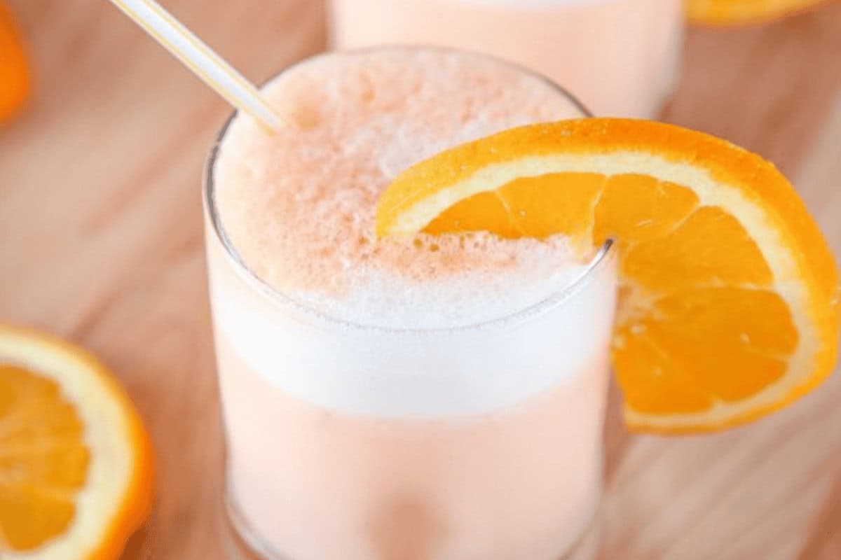 A glass of frothy orange drink with a white straw and an orange slice garnish, placed on a wooden surface with additional orange slices nearby.