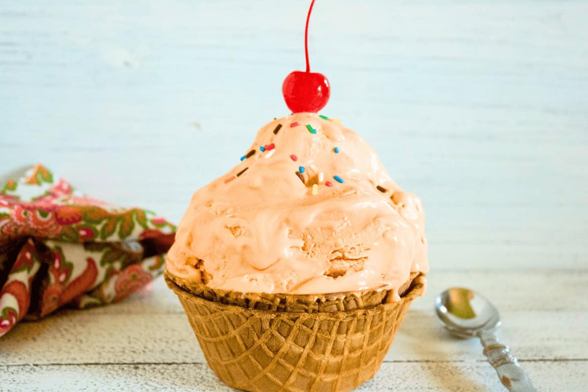 A scoop of orange ice cream with sprinkles and a cherry on top in a waffle bowl, with a spoon and patterned napkin beside it.