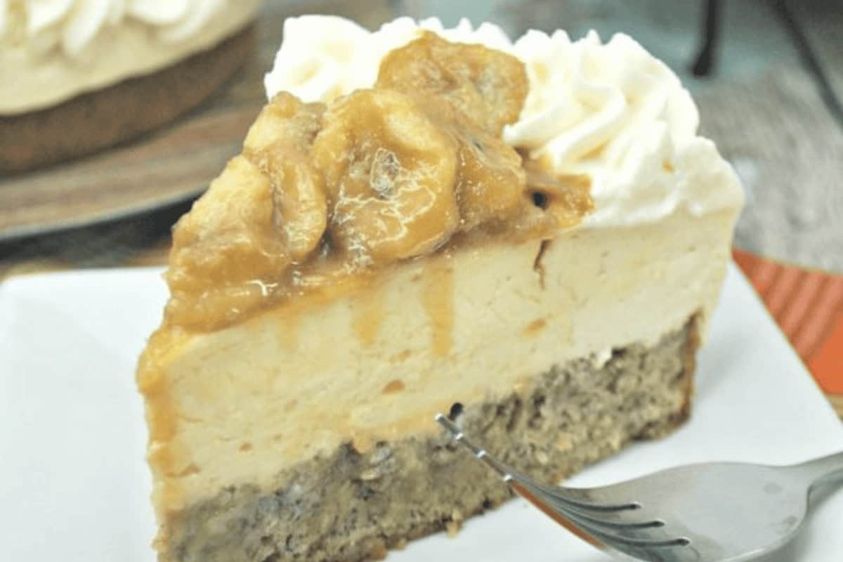 A slice of layered banana cheesecake topped with caramelized banana slices and whipped cream on a white plate, with a fork in front.
