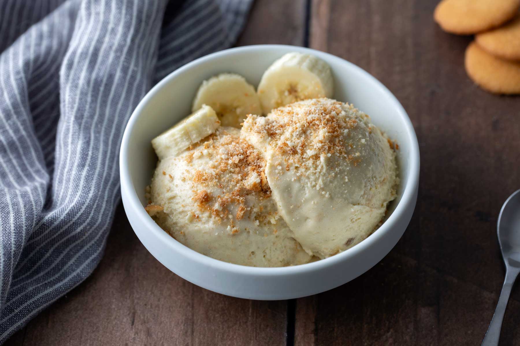 A white bowl with two scoops of ice cream, banana slices, and cookie crumbs on top sits on a wooden surface next to a striped cloth and cookies.