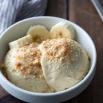 A white bowl with two scoops of ice cream topped with crumbled nuts, garnished with banana slices, on a wooden table next to a spoon and a striped cloth.