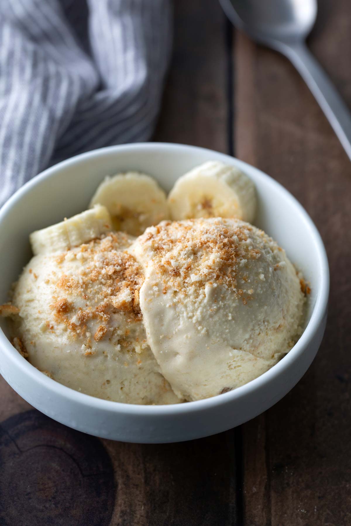 A white bowl with two scoops of ice cream topped with crumbled nuts, garnished with banana slices, on a wooden table next to a spoon and a striped cloth.