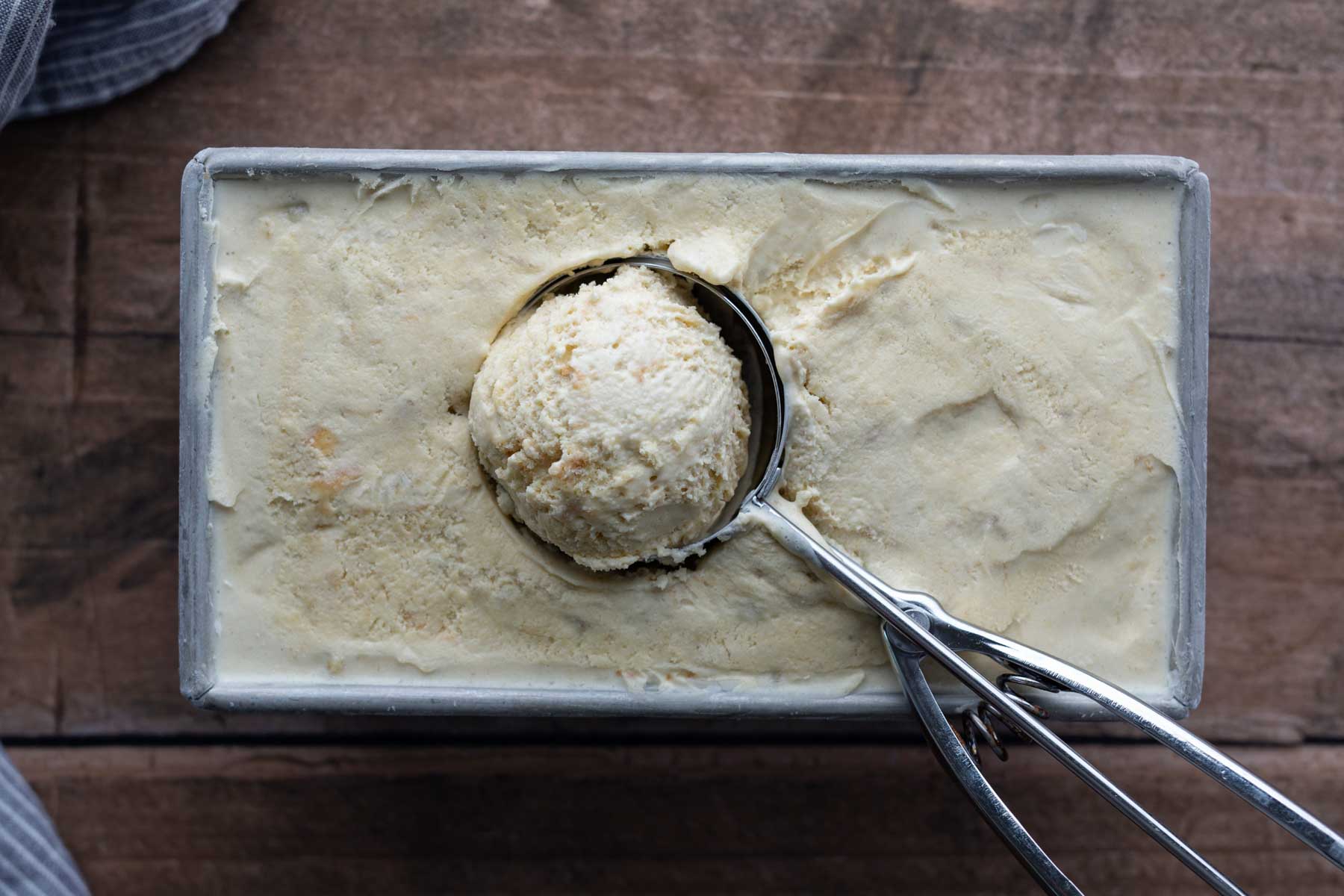 A metal ice cream scoop holding a round scoop of vanilla ice cream rests in a rectangular container filled with more ice cream on a wooden surface.