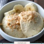 A white bowl contains scoops of banana pudding ice cream, topped with crushed cookies and banana slices.