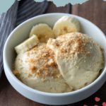 A white bowl of banana pudding ice cream topped with cookie crumbs and banana slices, placed on a wooden surface.