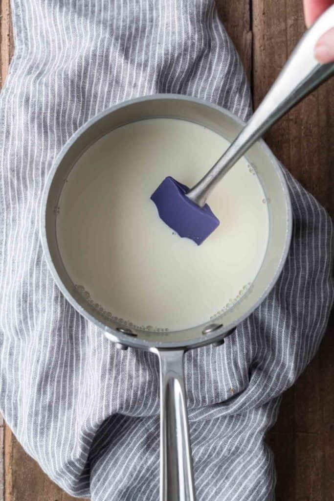 A saucepan filled with milk is being stirred with a blue spatula, placed on a gray and white striped towel on a wooden surface.
