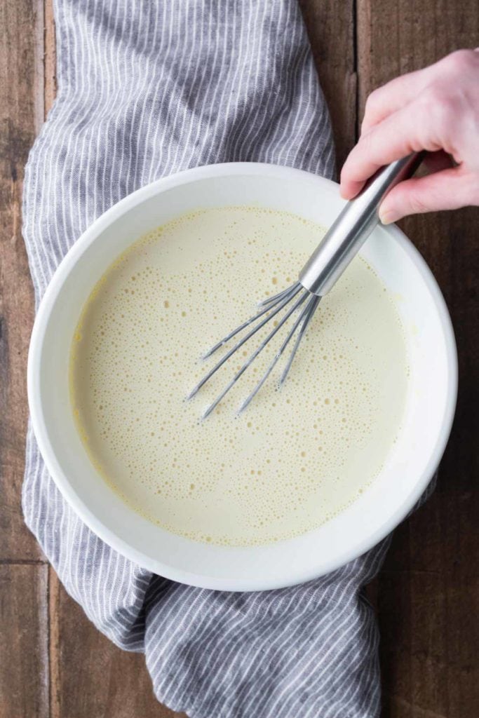 A hand whisking a bowl of pale yellow batter on a wooden surface with a gray striped cloth underneath.