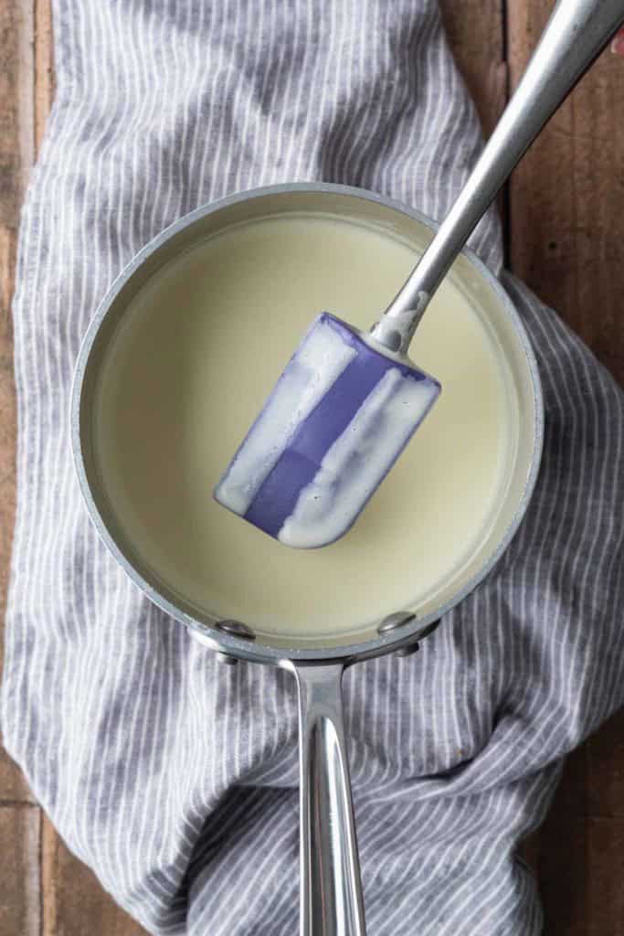A saucepan filled with a light-colored liquid sits on a striped cloth, with a spatula resting inside the pan.